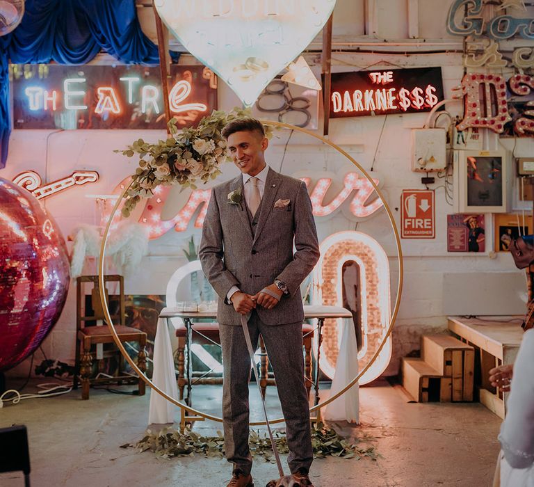 Groom in grey three piece wedding suit with yellow suit and yellow pocket square waiting at the end of the alter with puppy and numerous bright neon signs, LED signs and large wooden circular arch with rose decorations attached 