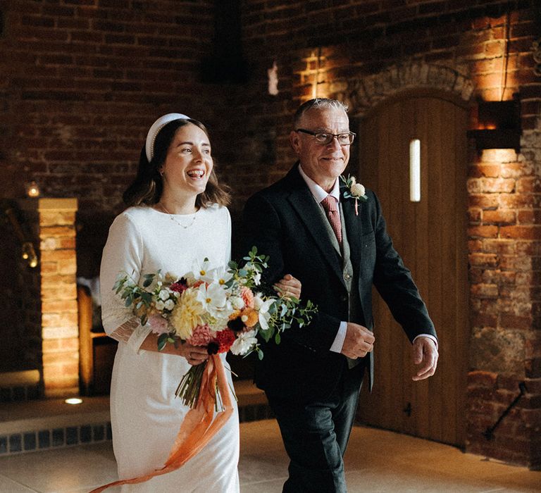 Father of the bride in three piece suit wearing a white flower buttonhole smiles as he walks the bride in a high neck three quarter length sleeve down the aisle 