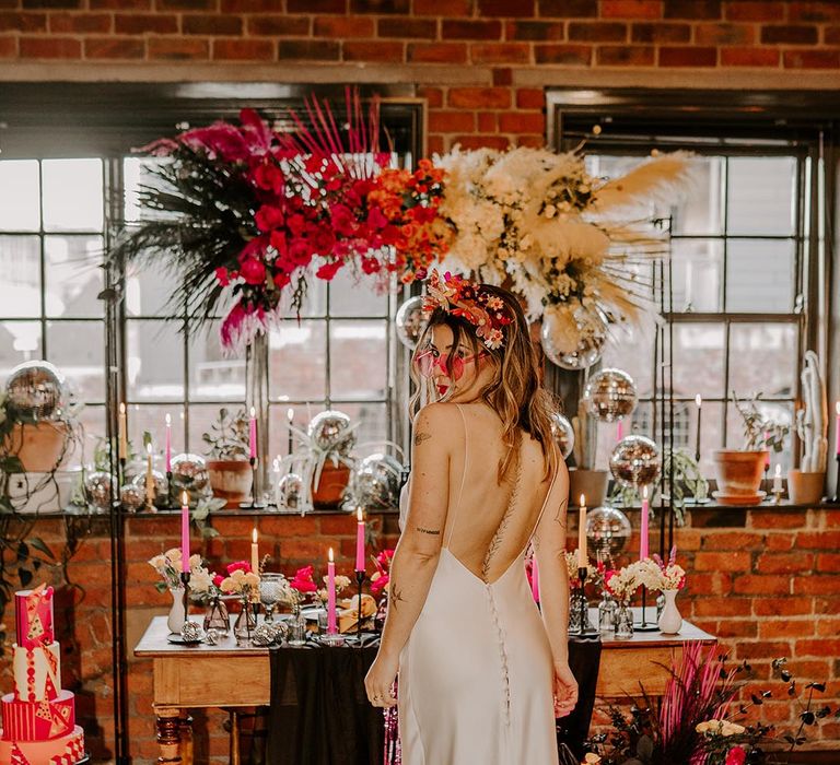 Bride in low back detail satin wedding dress and sheer pink sunglasses with pink floral bouquet standing in front of wedding tablescape with pink metallic streamers, hanging disco ball decorations, hot pink neon wedding sign, yellow smiley face neon sign, viva magenta, fuchsia and hot pink tapered candles and suspended pink and yellow rose flowers with foliage 