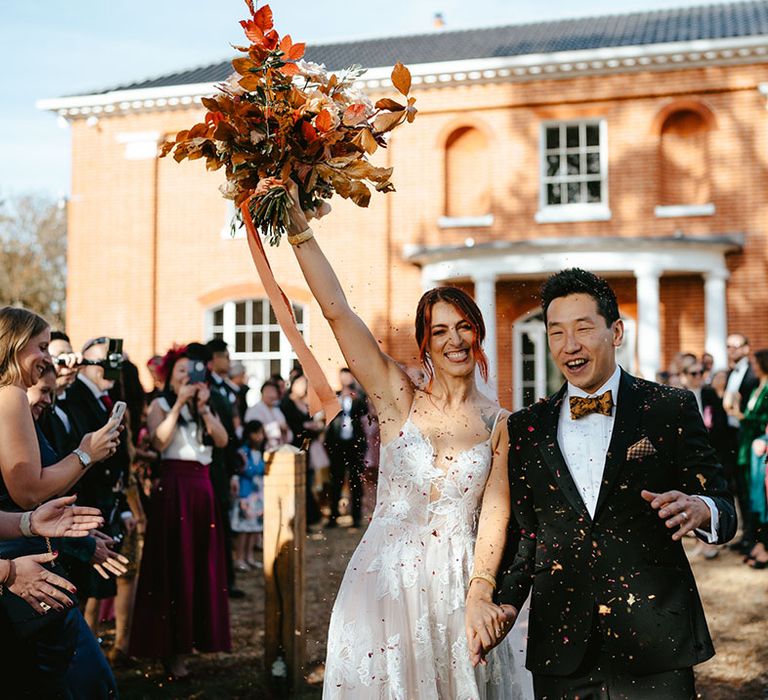 Bride lifts burnt orange bridal bouquet into the air as she walks through confetti exit with her groom 