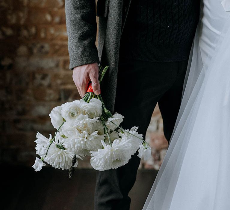 Bride standing with groom holding bouquet with white roses, white peonies and white chrysanthemums tied with red ribbon 