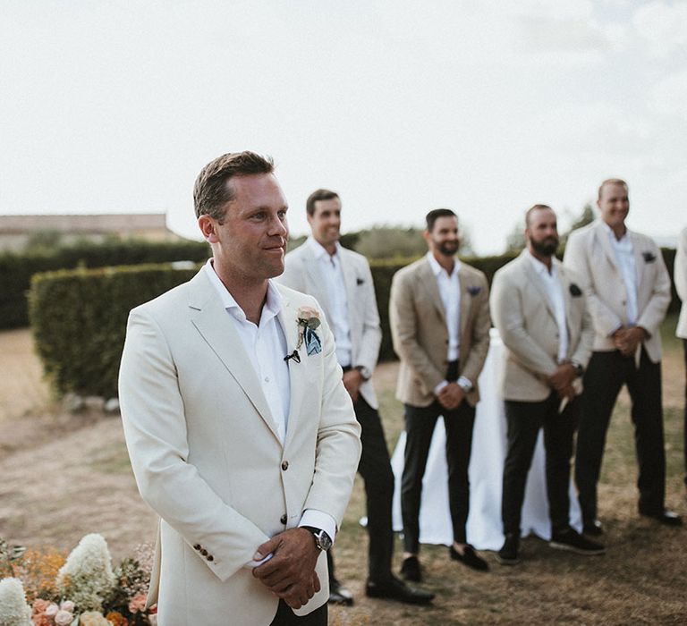 Groom wears cream coloured suit jacket with white shirt and floral buttonhole