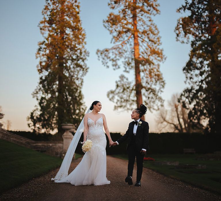 Bride in lace sleeveless v neck Enzoani wedding dress with tulle finish and church length veil walking hand in hand with bride in black snakeskin print tuxedo with black bowtie and white rose boutonniere walking through the grounds of Grittleton House wedding venue 