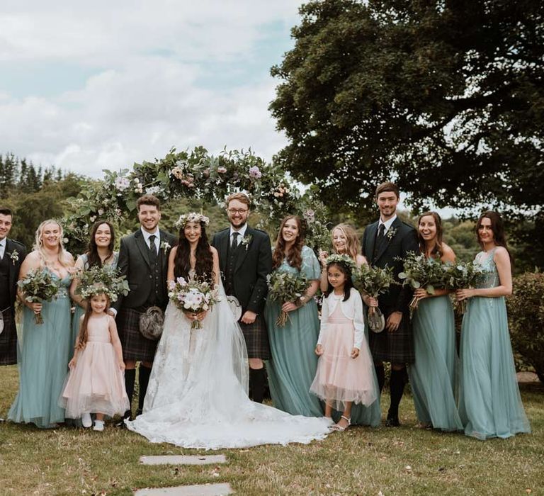 Groom in dark grey blazer and waistcoat, dark tartan tie with white flower boutonniere, dark grey and blue tartan kilt and wedding sporran, bride in ethereal, celestial 3d appliqué sheer overlay wedding dress and bridal flower crown holding large white rose, peony, dried flower and foliage bridal bouquet, bridesmaids in mismatched embellished teal bridesmaid dresses, groomsmen in classic wedding kilts and flower girl in baby pink dress with white cardigan and flower crown 