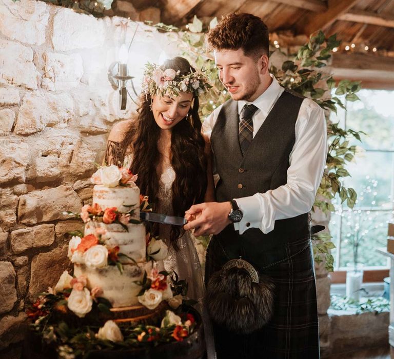 Bride cutting the three-tiered buttercream wedding cake with white garden rose and seasonal fruit cake decor with groom in dark grey waistcoat and tartan tie and kilt at The Barn at Harburn wedding venue 