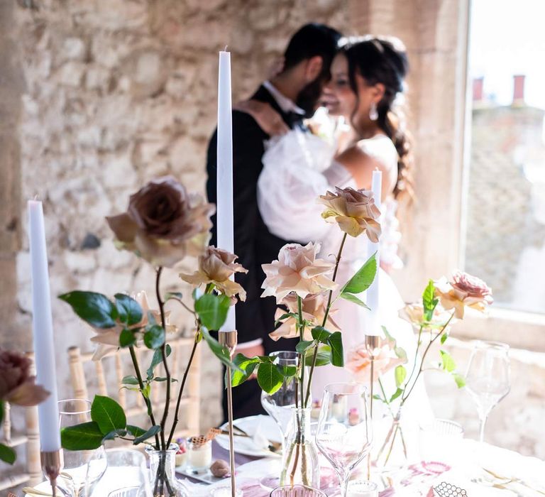 Bride and groom embracing in front of contemporary wedding tablescape with lavender tablecloth, lilac tulle table runner, blush garden rose centrepieces, tea light candles and tapered candles 