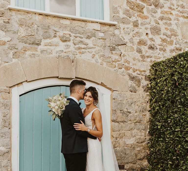 Light blue colourful door with the bride in a fitted beaded wedding dress embracing the groom in a navy and black tuxedo 