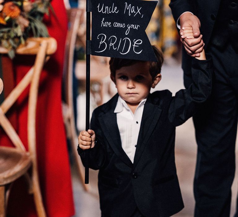 Page boy in white shirt and black trousers and blazer with black and white sign reading 'uncle Max, here comes your bride' 