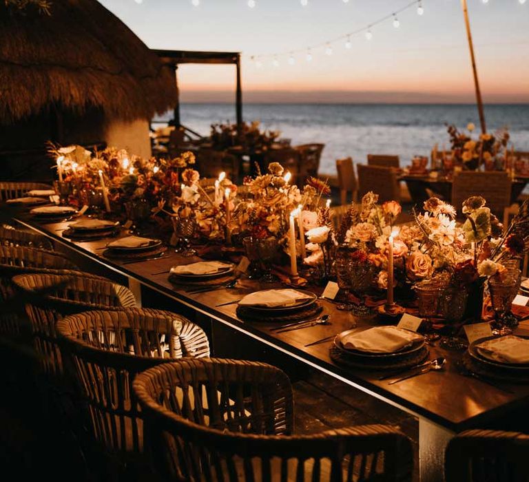 Rustic wedding tablescape by the Mexican sea with wicker chairs, burnt orange dried roses, dried flower and foliage floral centrepieces, wooden slate wedding table numbers, light orange tapered candles and burnt orange table runners under suspended fairy light wedding lighting 