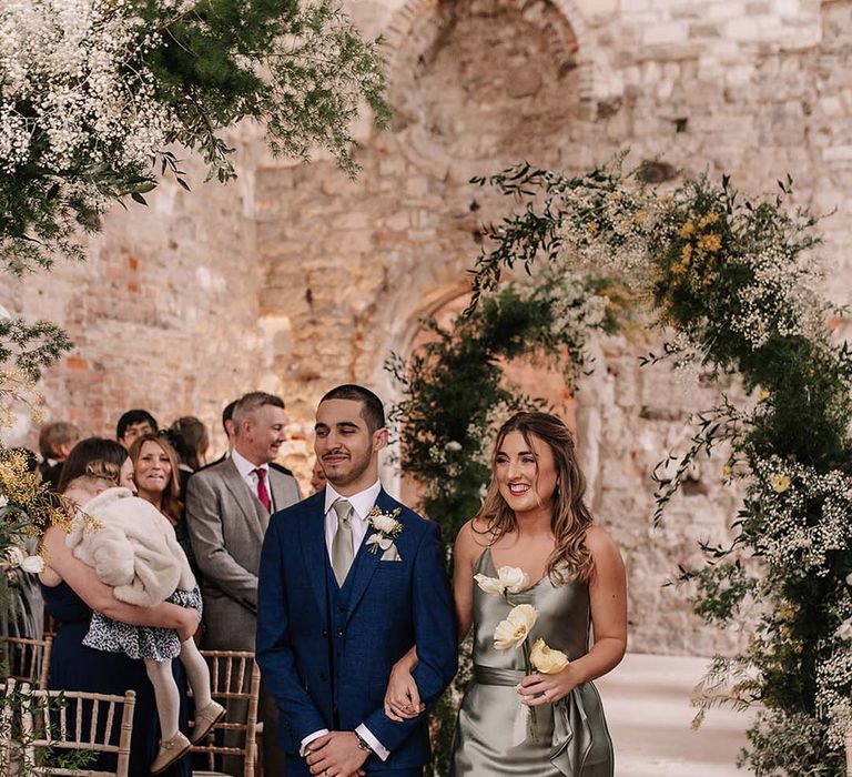 Bridesmaids in green mismatched bridesmaid dresses walk down the aisle accompanied by a groomsman 