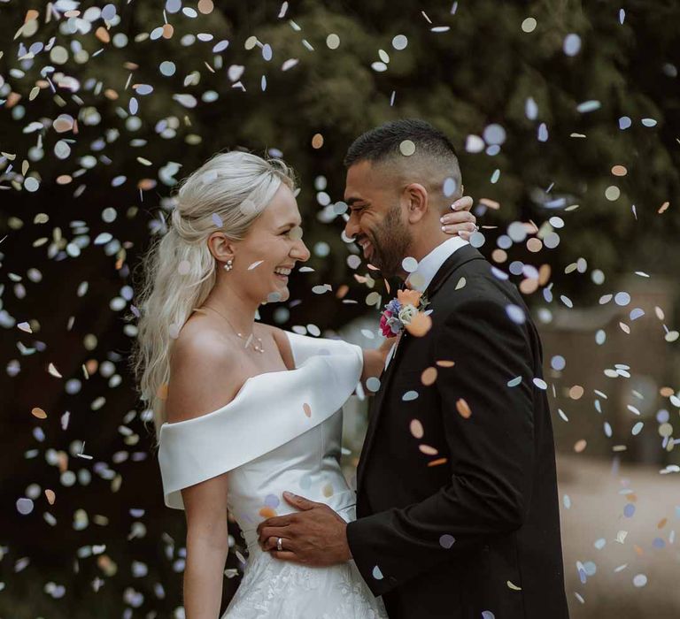 Groom in classic three piece grooms tuxedo with colourful spring boutonniere embracing bride in off the shoulder lace wedding dress holding colourful spring bridal bouquet doing colourful confetti photoshoot 