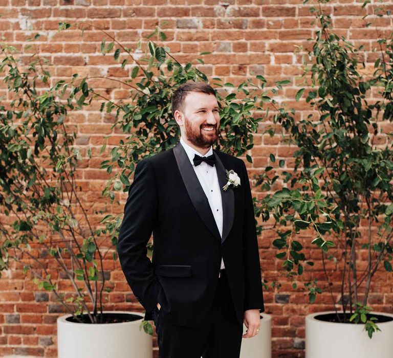 Groom standing in the courtyard of Brickhouse Vineyard Devon wedding venue in classic black grooms three piece tuxedo with white rose boutonniere and black bowtie 