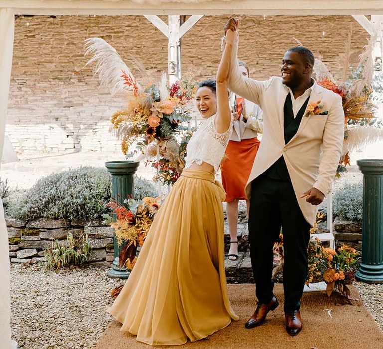 Bride in white mesh lace bridal crop top and yellow wedding skirt holding hands and cheering with groom in cream morning suit with forest green grooms waistcoat and trousers, forest green pocket square and orange boutonniere 