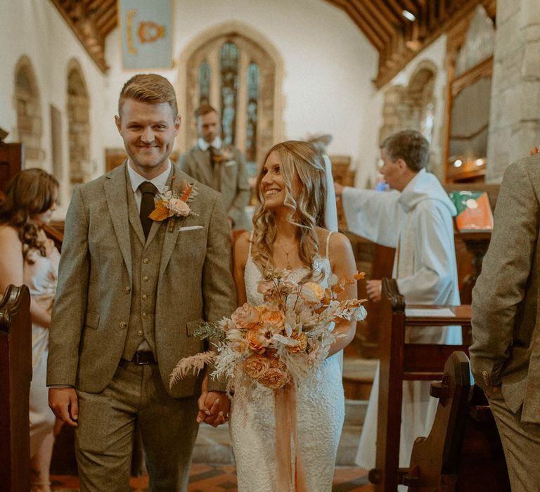 Groom in three piece grey beige wedding suit walking back down the aisle with the bride in a lace wedding dress holding orange autumnal bouquet with dried flowers