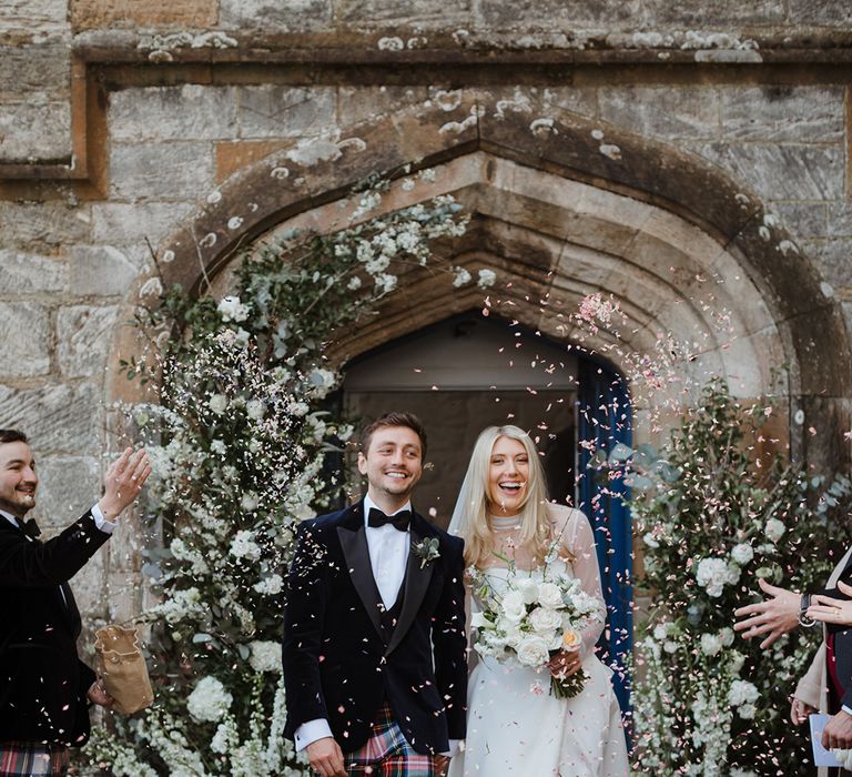 White wedding flower columns decorating the church entrance with the groom in tartan trousers and bride in mesh long sleeve wedding dress as guests throw confetti