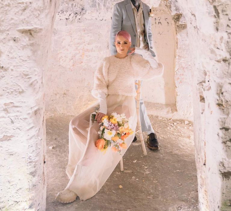 Bride in sheer overlay wedding dress and fluffy bridal coverup holding colourful bridal bouquet standing with groom in baby blue linen suit, personalised embroidered linen shirt and garden rose and pampas grass boutonniere in Margate Caves