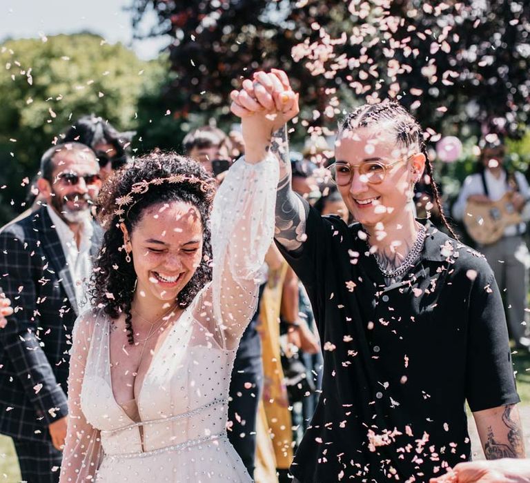 Bride in silver chains, black shirt and black trousers doing colourful confetti exit after handfasting ceremony with bride in long sleeve v-neck sheer polka dot overlay sparkly wedding dress and gold celestial bridal headband 