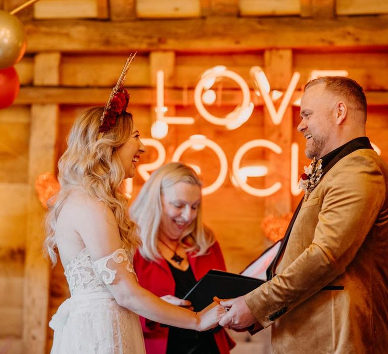 Bride in off shoulder lace wedding dress with corset style top and red rose and gold halo bridal crown holding hands with groom in crushed velvet burnt orange grooms blazer and dried flower boutonniere standing in front of large wedding neon sign at Southlands Barn