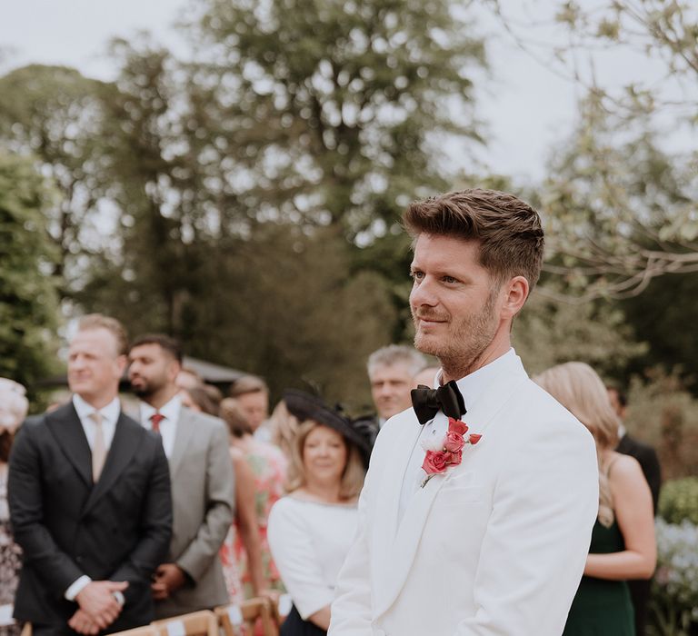 Groom wearing a white tux for the outdoor wedding with a red and pink wedding buttonhole 