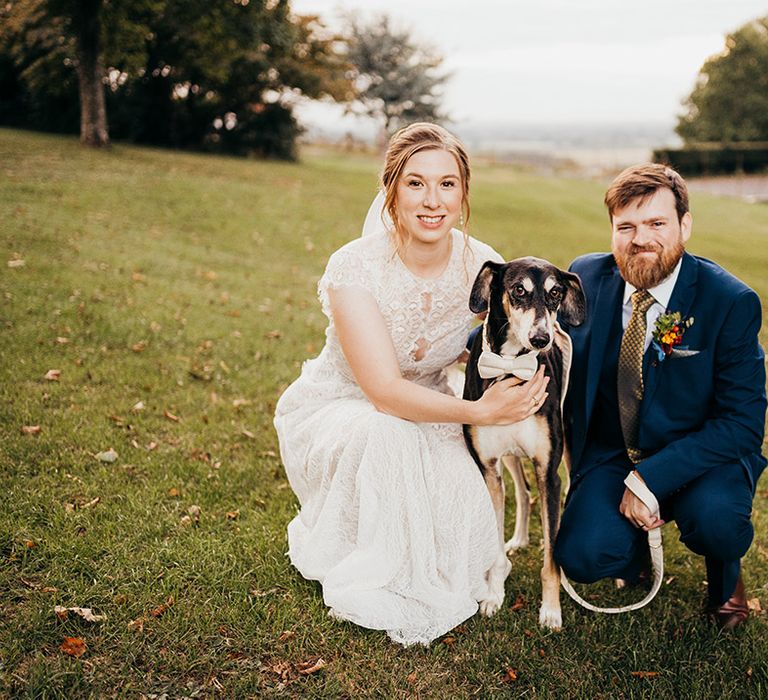 Dog wearing white bow tie standing in between bride in lace wedding dress with groom in blue suit 