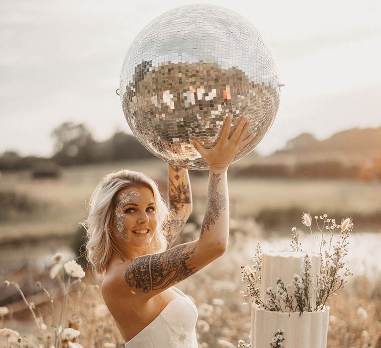 Bride with tattoos in strapless wedding dress waving giant disco ball in the air 