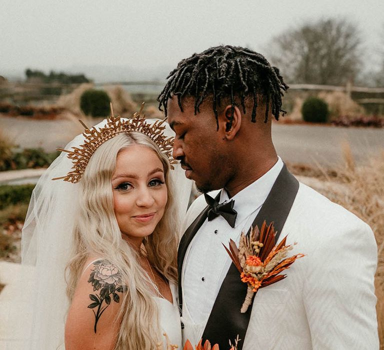 Groom in white tuxedo jacket with bride wearing gold crown holding dried wedding flower bouquet 