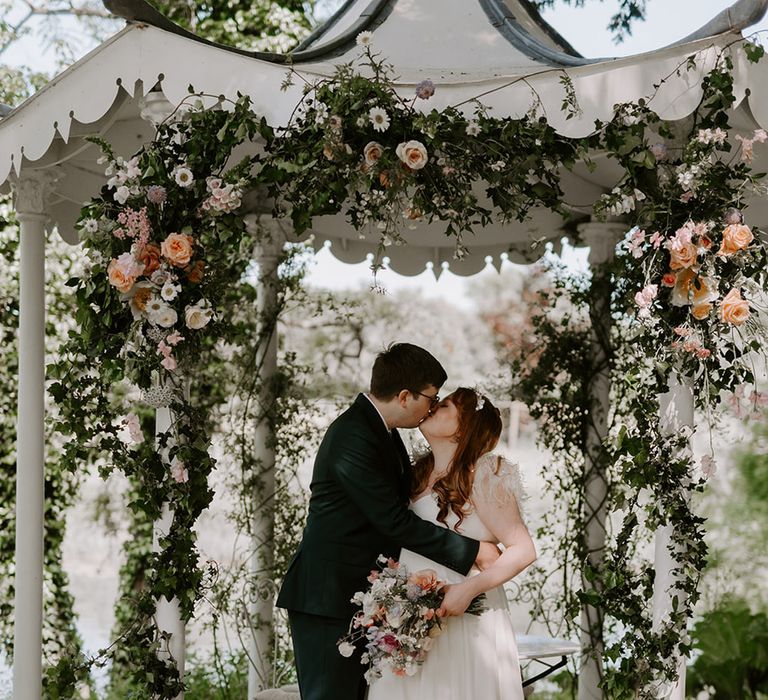 The bride and groom share a kiss under the outdoor gazebo surrounded by neutral wedding floral arrangements 