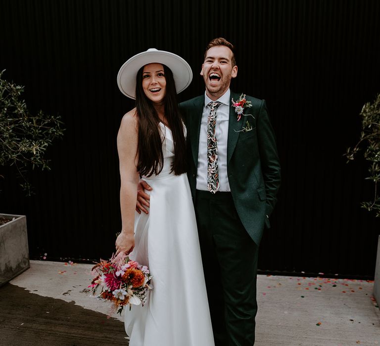 Bride in Suzanne Neville wedding dress with white pearl bridal hat with groom in dark green suit 