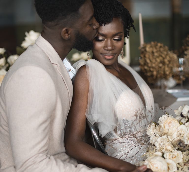 Groom in beige suit kissing his brides forehead with naturally curly hair and smokey eye makeup 