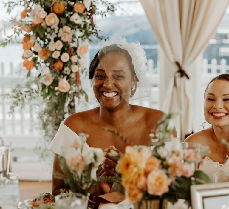 Two brides smiling during their wedding reception whilst sitting at their sweetheart table 