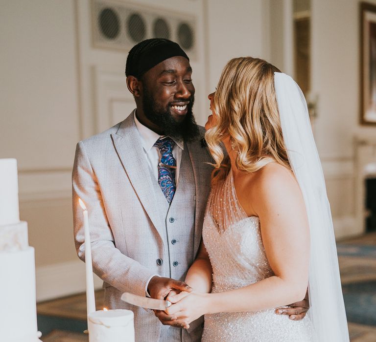 Black groom in a grey check suit and his bride in a sparkly one shoulder wedding dress cutting the marble wedding cake 