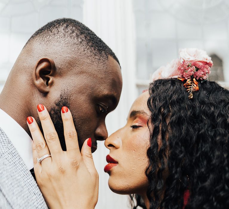 A bride and groom touch noses for wedding portrait session. She has loose curly hair and wears flowers in her hair. She has red lipstick and red nails. He has skin fade and wears a grey suit.