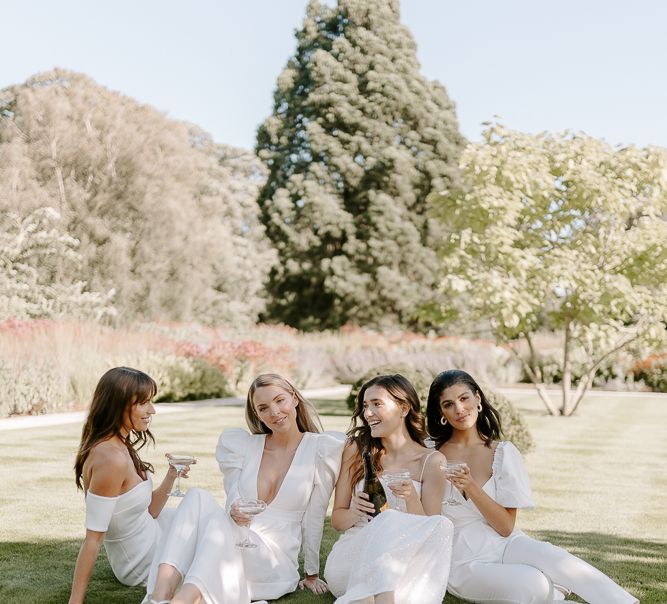 Bridal party in white dresses, sandals and boots sitting on the lawn at Middleton Lodge drinking champagne 