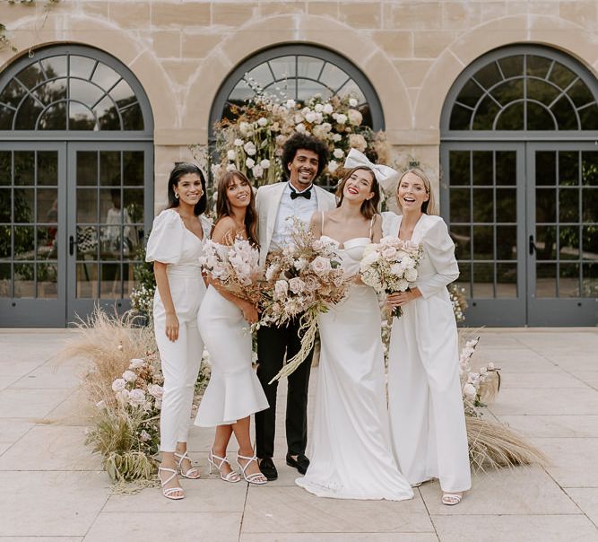 Bridesmaids in all white dresses and jumpsuits standing with the bride and groom in a bow and tuxedo jacket at The Fig House, Middleton Lodge