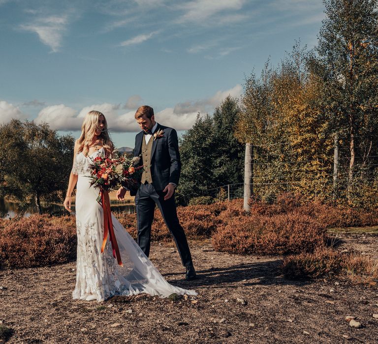 Bride walks in appliqué wedding gown carrying Autumnal bouquet tied with orange ribbon  