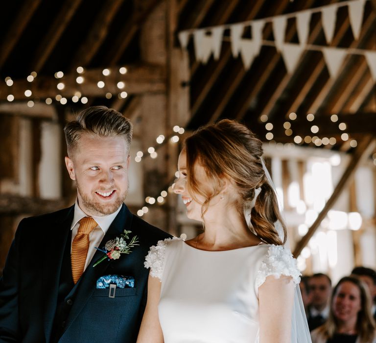 Groom in a navy suit with orange tie, blue paisley pocket square and succulent buttonhole smiling at his bride at the altar 