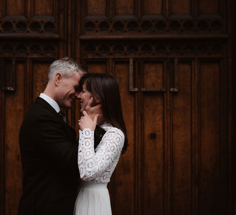 Smiling groom in brown woollen blazer holds face of bride in long sleeved lace top Self Portrait wedding dress in front of wooden door in Bristol