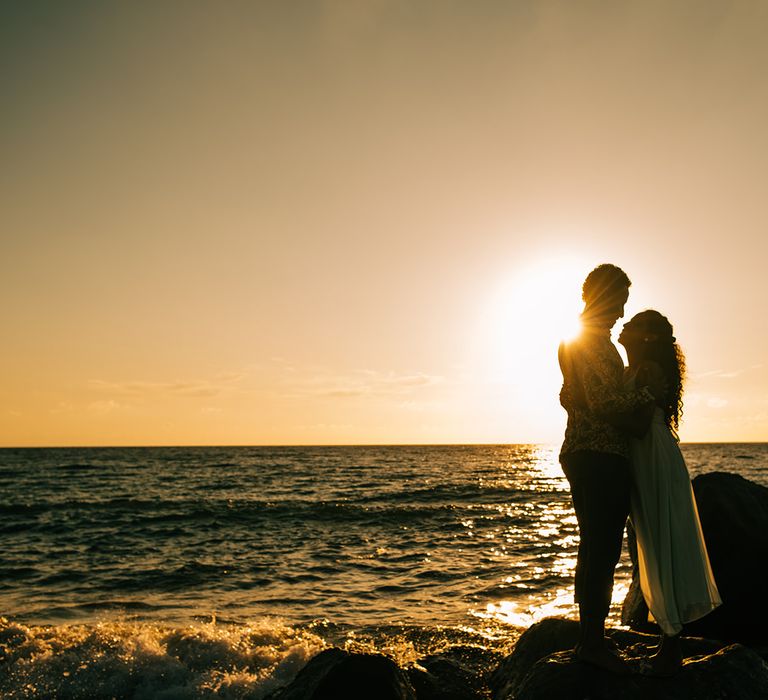 Bride & groom stand in front of the sun setting on the beach