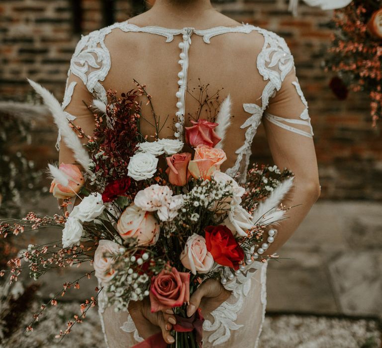 Bride with curly wedding hair up-do with pearl hair accessories, wearing a mesh and embroidered button back long sleeve dress, holding a bouquet of red, white and neutral flowers