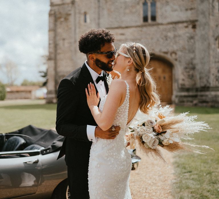 Groom in a tuxedo kissing his bride in an appliue wedding dress with sleek ponytail and dried flower bouquet outside Pentney Abbey, Norfolk