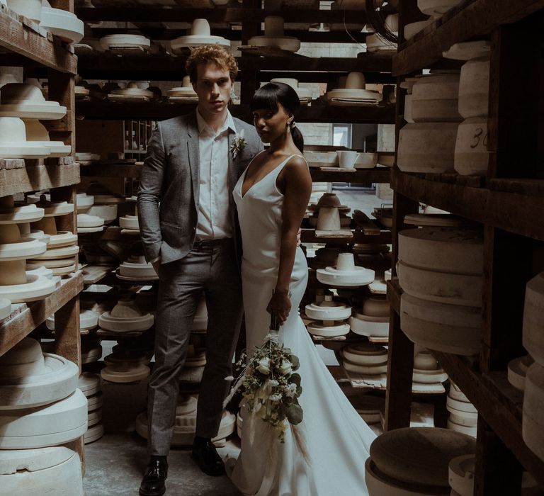 A bride and groom stand in front shelves of ceramics. She holds a white dried flower bouquet down to her side for a simple elegant wedding. 