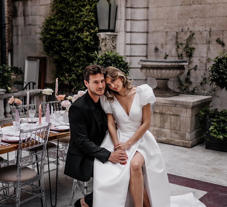 Bride in a puff sleeve wedding dress with front split sitting on her grooms lap at their stylish outdoor wedding reception 