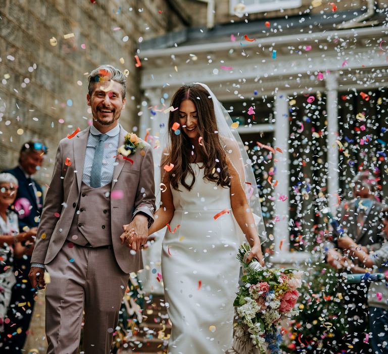 Bride in white strapless Rebecca Vallance Dress and veil holding multicoloured bouquet walks hand in hand with groom in brown Moss Bros suit and blue tie as they walk through multicoloured confetti after Harrogate wedding