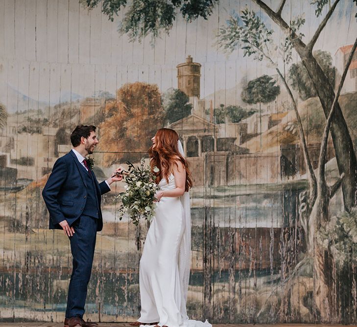 Groom in a navy suit and bride in a slip wedding dress laughing on the stage at The Larmer Tree wedding venue's outdoor theatre 