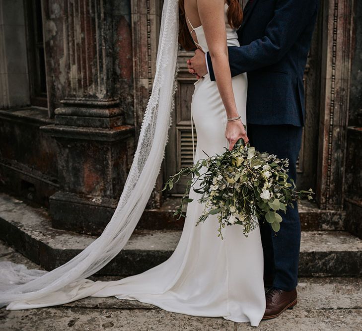 Stylish bride in a fitted wedding dress with delicate lace cathedral length veil holding her green and white bouquet by her side 