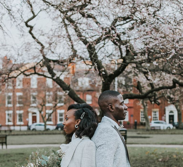 A Black couple stand back to back in a park for a wedding portrait. He wears a white tux jacket and she wears her hair clipped up tone side with a white blazer jacket. 