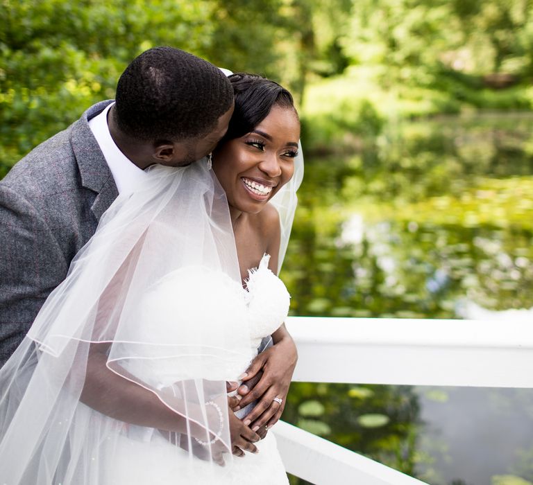 Groom kisses bride on the cheek as they stand outside in picturesque setting