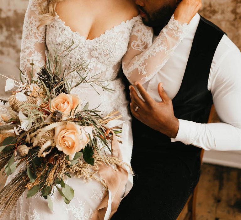 Bride sitting on the groom's lap as he faces into her shoulder and neck. She is holding a bouquet of peach coloured roses and wild grasses