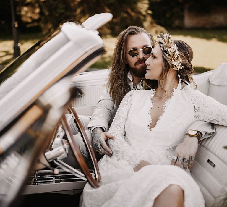 Boho bride in a lace wedding dress and dried flower crown and stylish groom in a beige suit and sunglasses sitting in a white mustang wedding car 