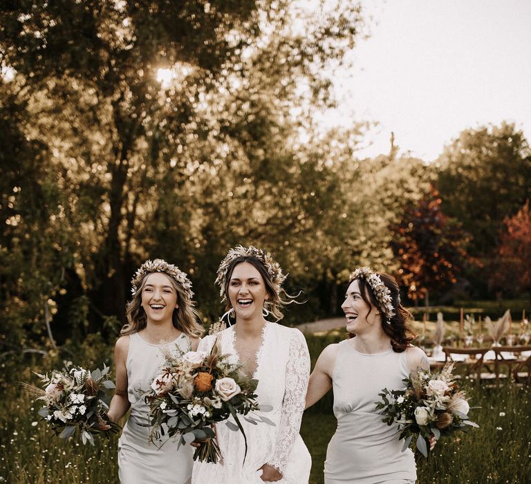 Bridal party portrait with bridesmaid in grey satin dresses and bride in a lace wedding dress with long sleeves. They all wear a dried flower crown and hold white and green bouquets 